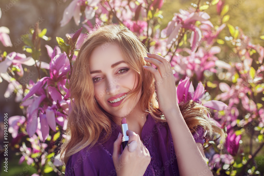 Fototapeta premium Outdoor portrait of a young beautiful happy smiling lady using lipstick near magnolia tree with flowers. Model looking at camera. Girl wearing stylish clothes. Female spring fashion