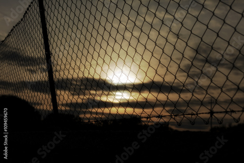 A fence covering the orange-colored sunset sky. Barbed wire on top. Symbolic shot: depression, jail, war.