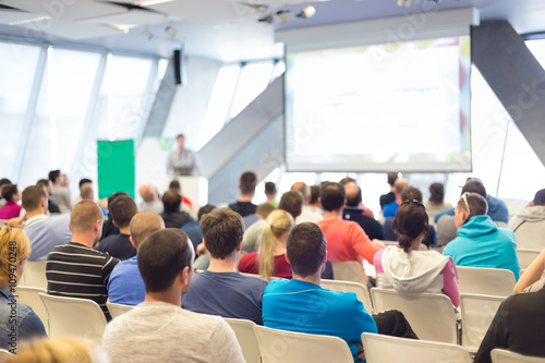 Man giving presentation in lecture hall. Male speeker having talk at public event. Participants listening to lecture. Rear view, focus on people in audience. photo