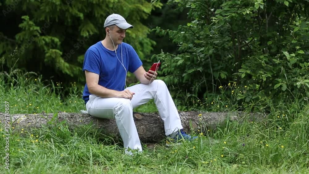 Man in dark blue t-shirt sits on a fallen tree in the forest and communicates via red smartphone. Adult man sits on a fallen tree in the forest and uses smartphone. Communication through skype Man in dark blue t-shirt sits on a fallen tree in the forest and communicates via red smartphone. Adult man sits on a fallen tree in the forest and uses smartphone. Communication through skype