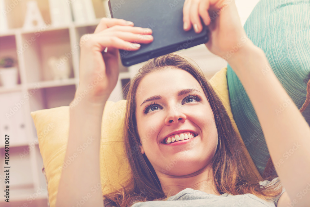 Naklejka premium Happy young woman reading an e-book on her couch