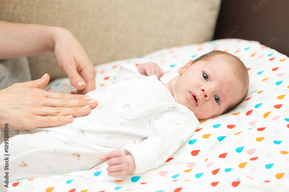 Newborn colicky baby dressed in white laying on her back, mother's hand on her tummy helping her with colic Newborn colicky baby dressed in white laying on her back, mother's hand on her tummy helping her with colic
