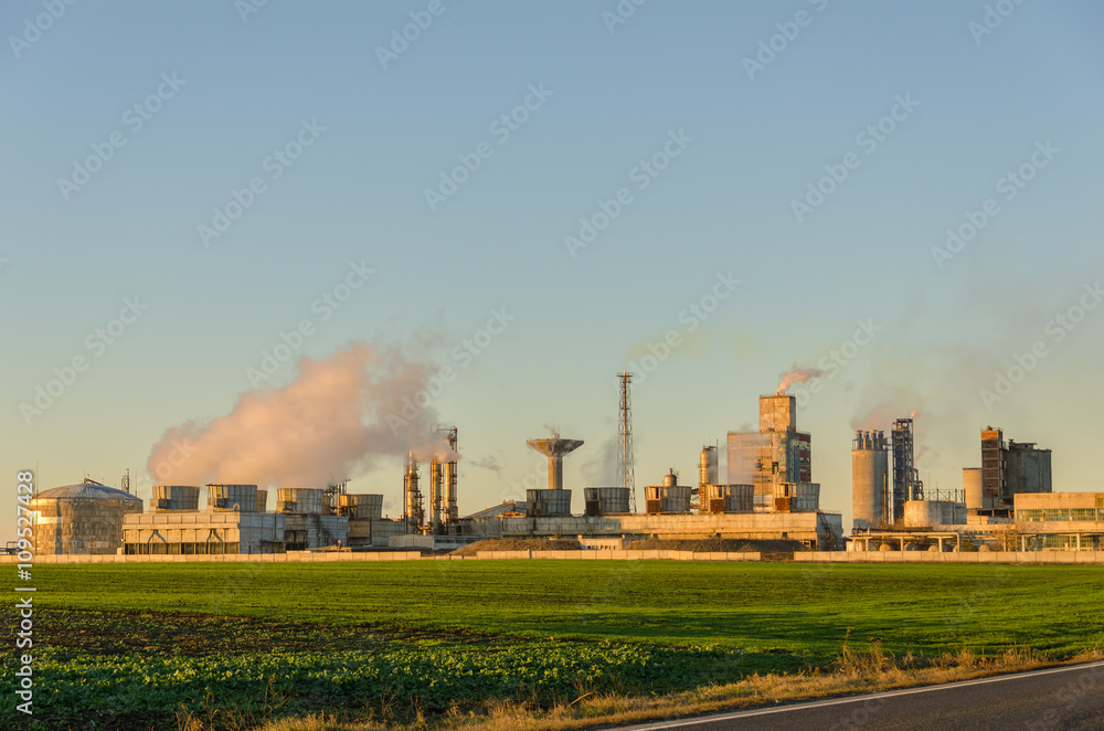 Chemical plant with smoke in sunset light on a green field against clear sky