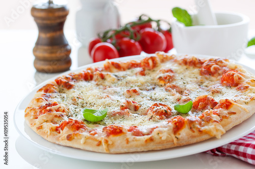 Fresh baked pizza with melted cheese, Italian herbs and tomato sauce and fresh basil on a white plate on a kitchen background, selective focus