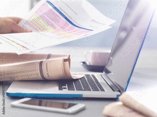 Man reviewing financial affairs using newspaper market data, investment statement and laptop