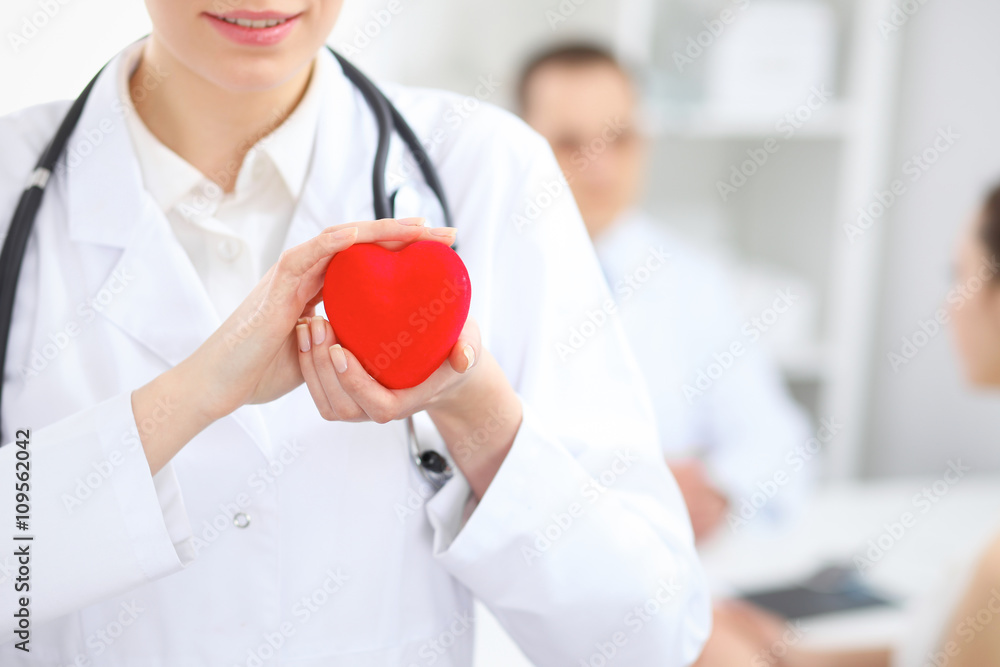 Female doctor with stethoscope holding heart.  Doctor and patient sitting in the background - obrazy, fototapety, plakaty 