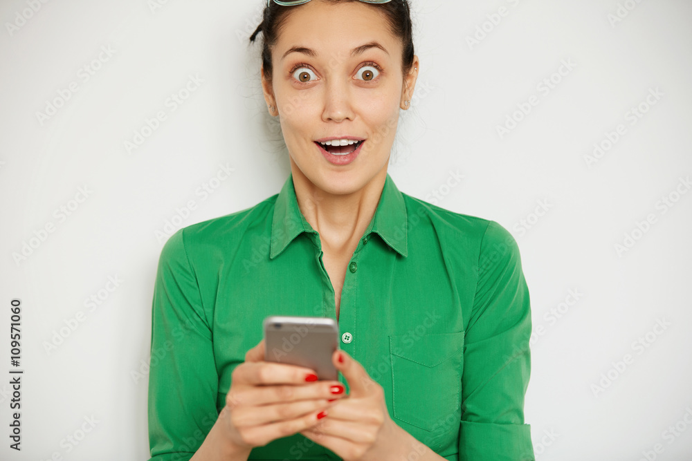 Human face expression and emotions. Cropped shot of surprised brunette woman holding cell phone and looking at the camera shocked with sale prices. Portrait of student girl astonished with good news Human face expression and emotions. Cropped shot of surprised brunette woman holding cell phone and looking at the camera shocked with sale prices. Portrait of student girl astonished with good news