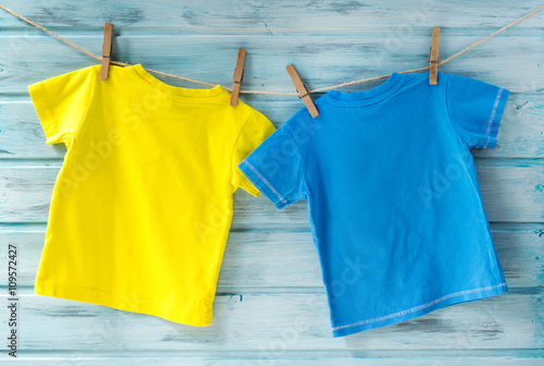 Two bright baby t-shirts hanging on a clothesline on a blue wooden background