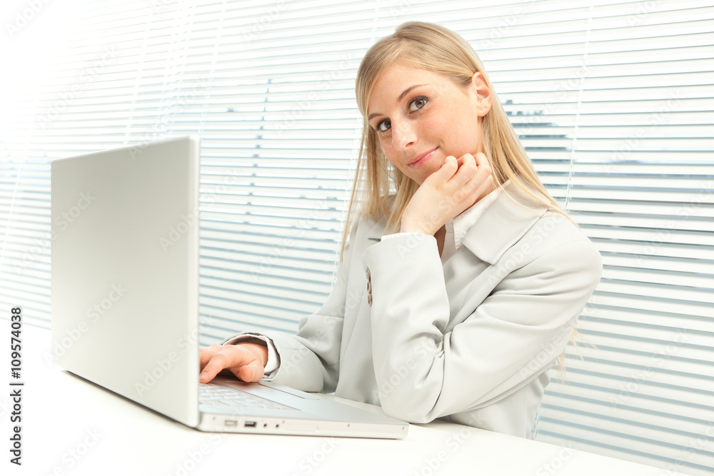 smiling blond businesswoman with laptop near venetian blind window smiling blond businesswoman with laptop near venetian blind window