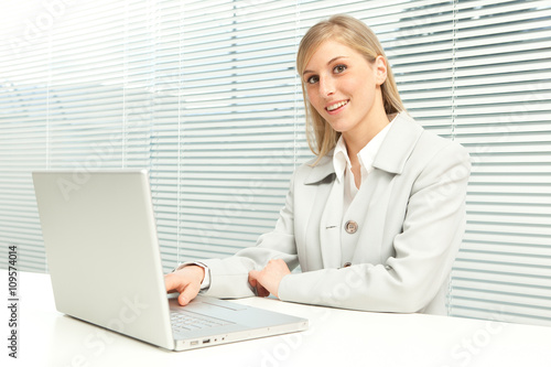 smiling blond businesswoman with laptop near venetian blind window