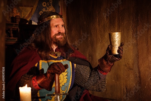 Old medieval king with goblet of wine on the throne in ancient castle interior.