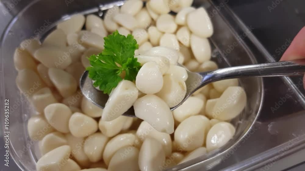 Metal spoon full of pickled garlic cloves, above a plastic container with more white cloves Metal spoon full of pickled garlic cloves, above a plastic container with more white cloves