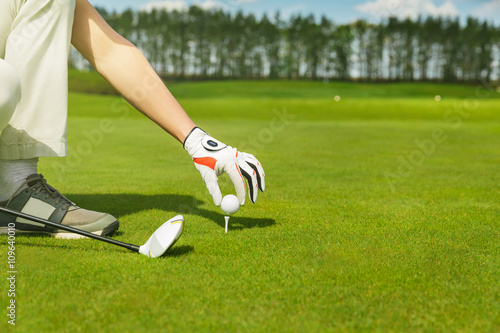 male hand in glove placing golf ball on tee over beautiful golf course with blue sky
