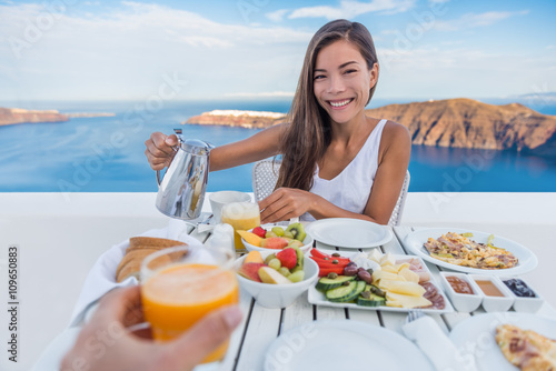 Couple eating breakfast. Smiling tourist woman drinking coffee and man drinking orange juice on terrace resort outdoor. Healthy and delicious food served for breakfast.  Santorini, Greece.
