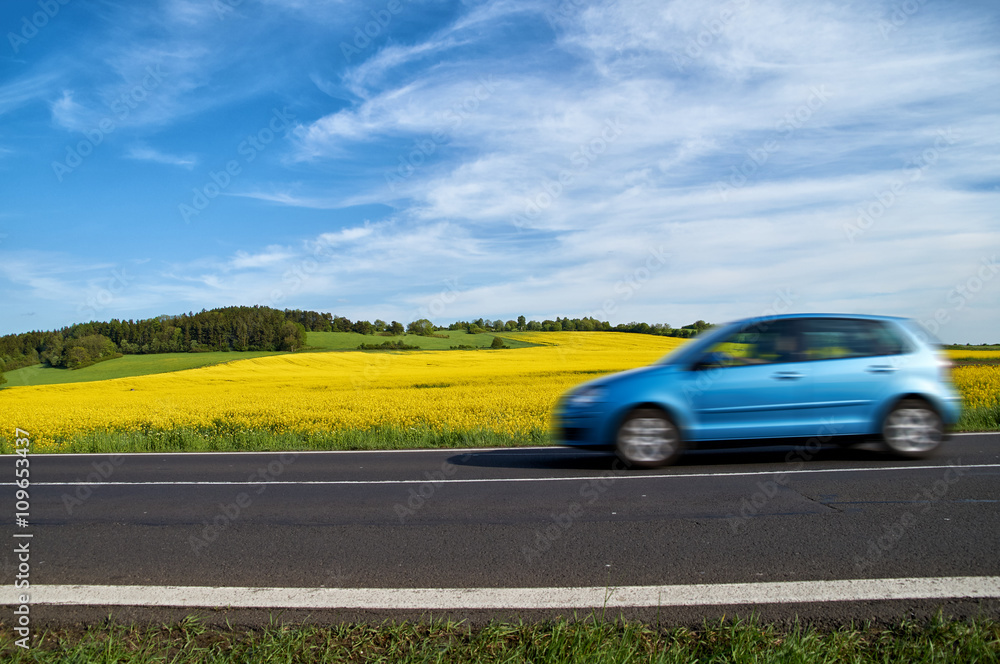 Motion blur blue passenger car speeding on the asphalt road around the yellow flowering rapeseed field. Sunny summer day.