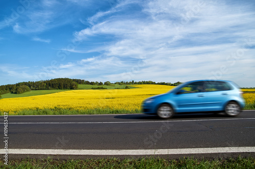 Motion blur blue passenger car speeding on the asphalt road around the yellow flowering rapeseed field. Sunny summer day.