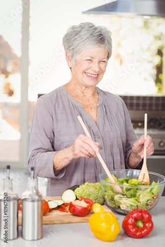 Happy senior woman tossing salad while standing in kitchen