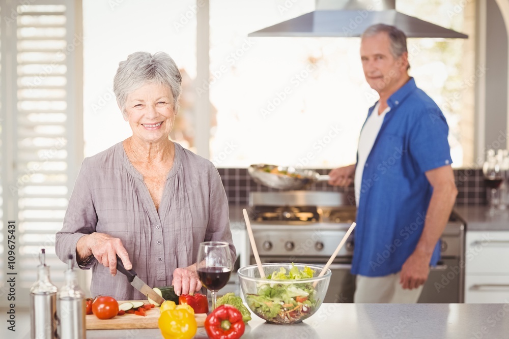 Happy senior woman cutting vegetables with husband in background Happy senior woman cutting vegetables with husband in background