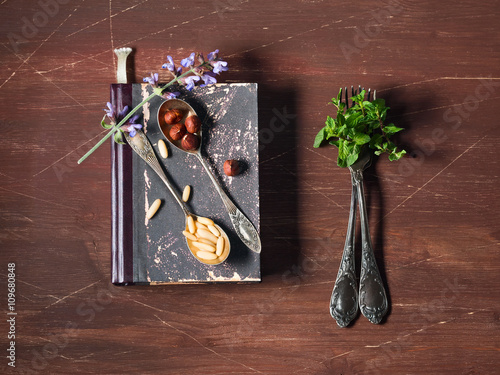 Vintage recipe book with nuts, flowers and mint leaves on vintage silver-plated forks, food still life on dark wooden background