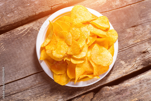 Potato chips pile on plate. Chips on brown wooden background. Fast snack at the diner. Example of processed food.