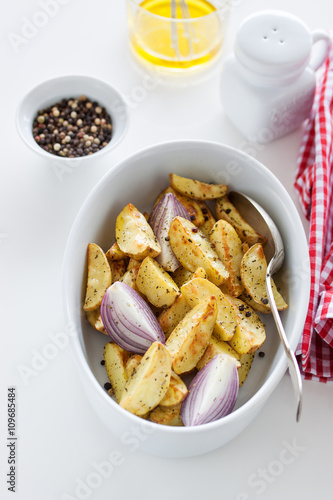 Oven-baked cut potato with red onion and Italian herbs, rustic, vintage or country style in a round bowl with white napkin on an kitchen background, top view