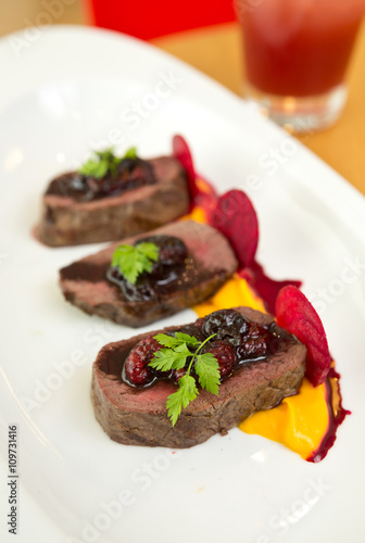 Organic Venison served with fresh raspberry raspberry sauce, beetroot slices and pumpkin puree, on a white plate.