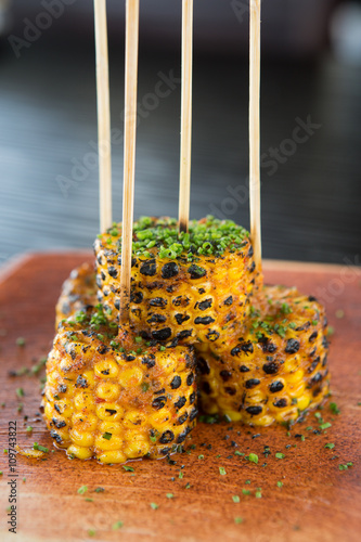 Grilled spicy sweetcorn on the cob, lightly covered with spices and chives, served on a wooden board and skewer. photo