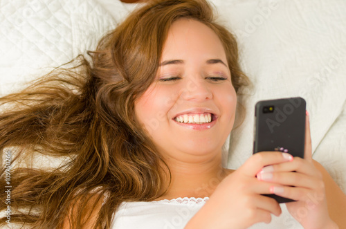 Young brunette woman in bed, comfortably resting on white sheets and pillow, holding mobile phone smiling