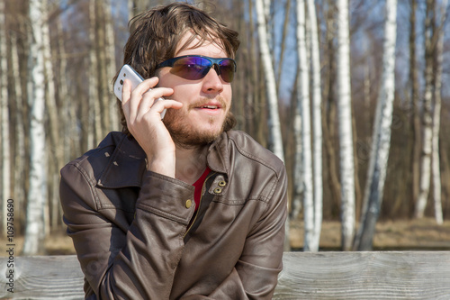 Young man wearing sunglasses, holding mobile phone, making a call and speaking on the phone at the park on the bench. Man in sunny spring day.