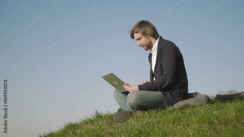 Young Man Talking On Skype Using a Laptop While Sitting On The Grass In The Park Young Man Talking On Skype Using a Laptop While Sitting On The Grass In The Park