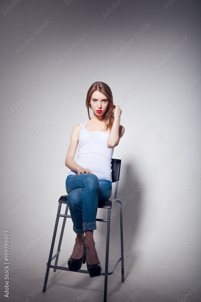Awesome caucasian attractive sexy professional female model with blond hair posing in studio wearing white shirt and black ragged jeans, sitting on chair, isolated on white background