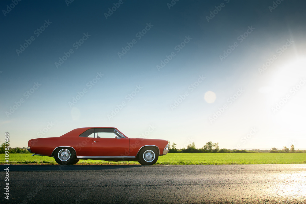 Obraz premium Retro czerwony samochód stojący na asfaltowej drodze o zachodzie słońca