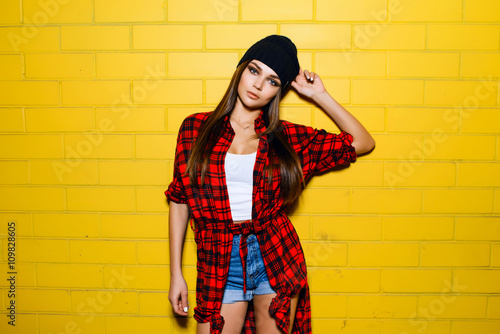 Beautiful young sexy hipster girl posing and smiling near urban yellow wall background in red plaid shirt, shorts, hat.