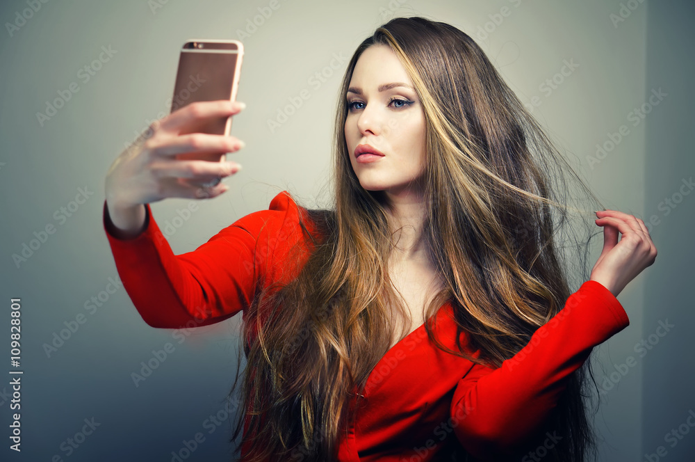 Naklejka premium Beautiful woman in red dress with perfect cute face and blue eyes taking selfie with smartphone in studio.