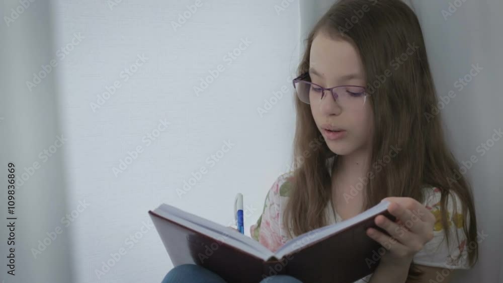 Beautiful little girl in glasses diligently does his homework at window. Beautiful little girl in glasses diligently does his homework at window.