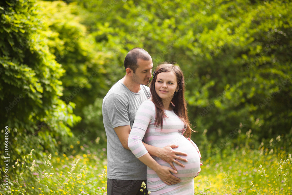 Fototapeta premium Happy couple waiting for baby. Man touches pregnant woman and hugs her belly outdoors in summer. New family concept. Paternity.