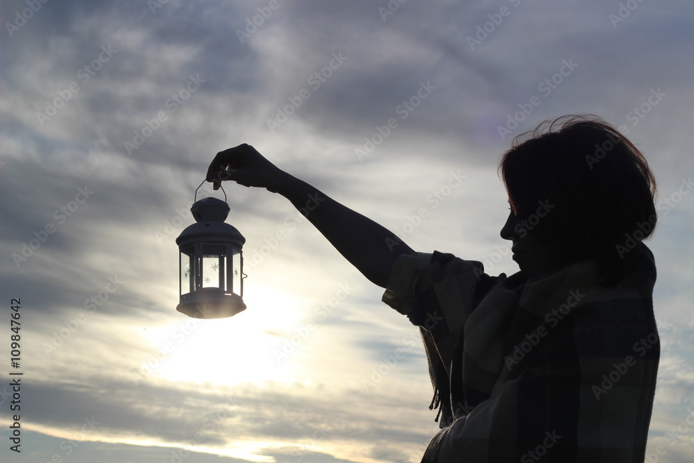 Girl silhouette with a candle lantern on backdrop of clouds and setting ...