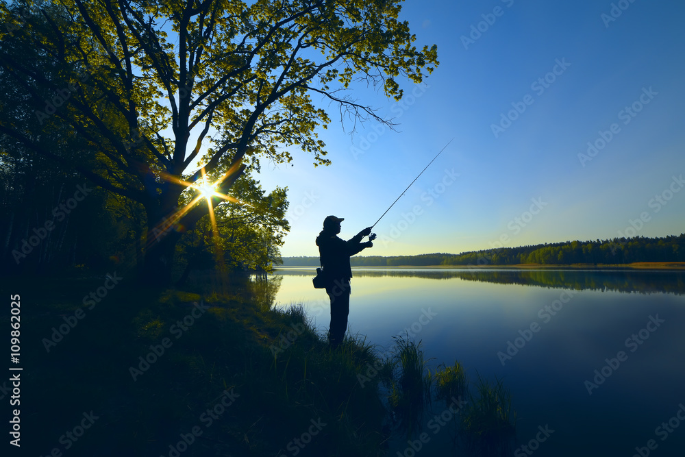 silhouette of a fisherman with a fishing rod on the shore of the lake, the river in the morning. The rays of the rising sun filtering through the tree leaves
