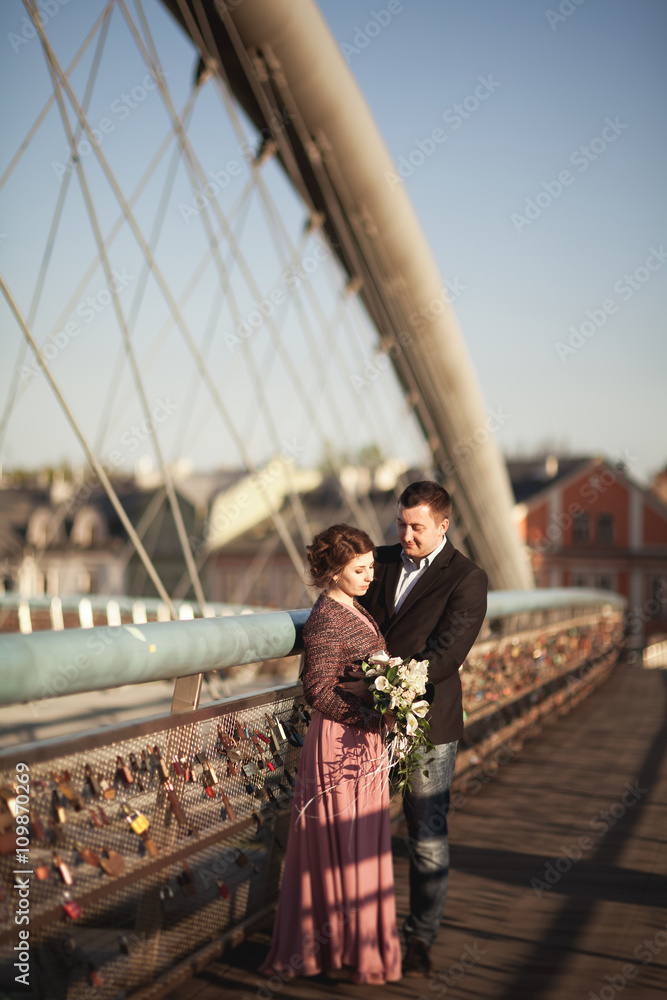 Fototapeta premium Stylish loving wedding couple, groom, bride with pink dress kissing and hugging on a bridge at sunset