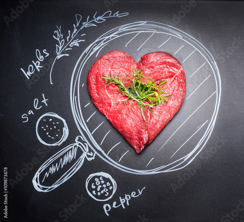 Heart shaped chop of meat on black chalkboard with  painted pan and ingredients, top view.For Meat lovers and eater photo