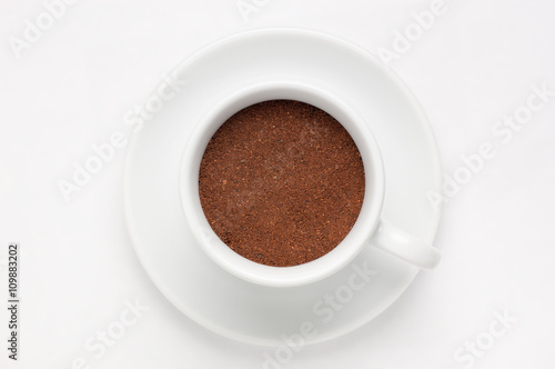 Coffee cup full of ground coffee on soucer against white background, top view photo