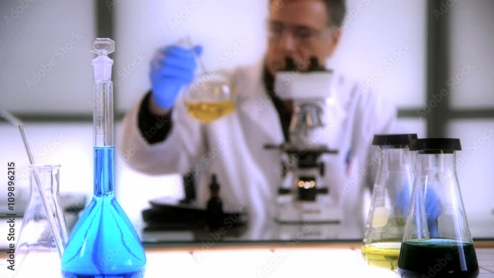 Focus remains on a volumetric flask and other lab glassware while a man working with a microscope works in the background. Focus remains on a volumetric flask and other lab glassware while a man working with a microscope works in the background.
