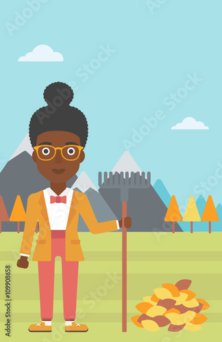 Woman with rake standing near heap of autumn leaves.