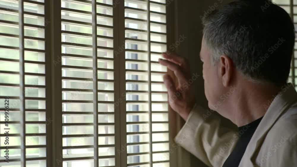 A mature man walks up to a window with shutters and smiles at what he sees outside. A mature man walks up to a window with shutters and smiles at what he sees outside.