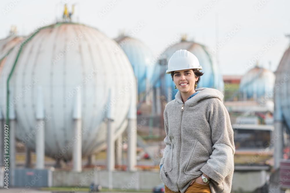 successful independent engineer smiling woman on industrial area with safety helmet looking to the camera. Pioneer woman at work with spherical tanks.