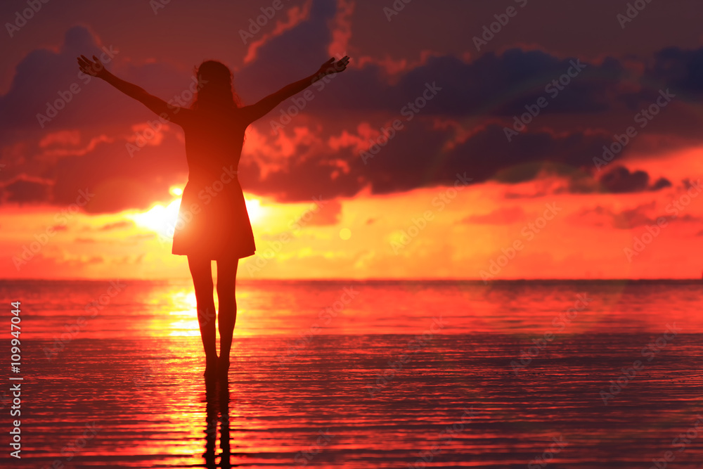 Silhouette of young woman standing on the sunset. Lady in dress raising her hands up in front of sun