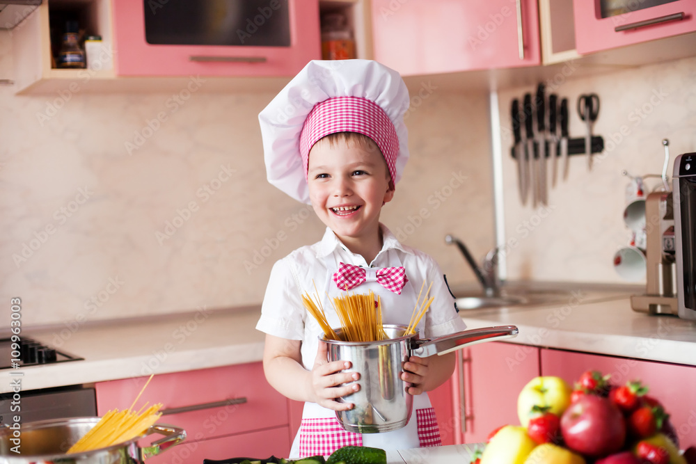 portrait of little boy in the hat of the chef and an apron. Little cooks chef in the kitchen preparing spaghetti.