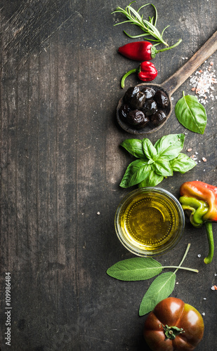 Food background with vegetables, herbs and condiment. Greek black olives, fresh basil, sage, rosemary, tomato, peppers, oil on dark rustic wooden background.
