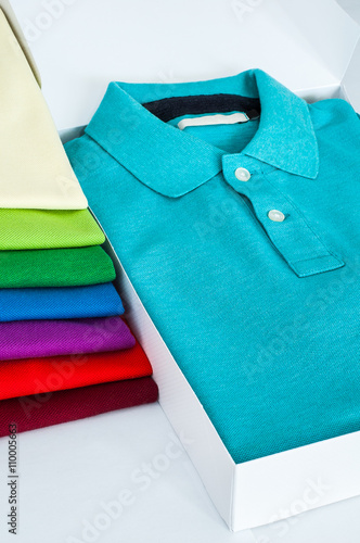 Luxurious fine material 100% cotton polo shirt displayed in a gift box with a pile of another polo shirts in many different colors.