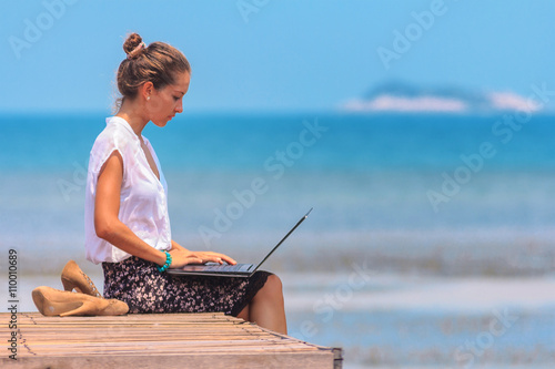Obraz na płótnie Young woman sitting with a laptop on the wooden platform edge at the sea. Lady freelancer in office suit working outdoor. w salonie